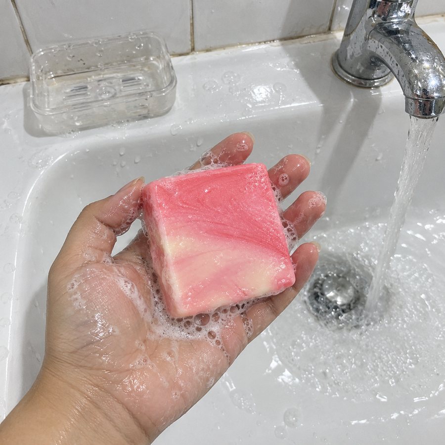 A wet hand holding the lathered marbled pink KIMIKO gluta bar over the sink